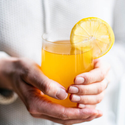 Woman in a white sweater holding a glass mug of yellow tea with a lemon slice.