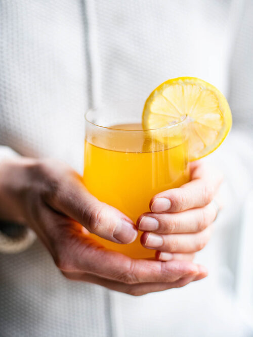 Woman in a white sweater holding a glass mug of yellow tea with a lemon slice.