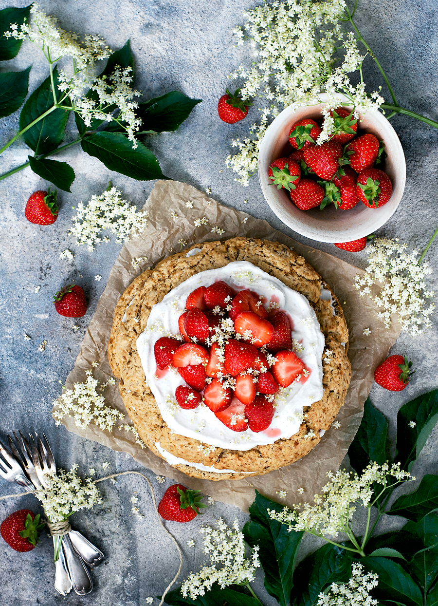 Giant scone cake layered with coconut whipped cream and strawberries, surrounded by elderflower, strawberries, and with a blue background