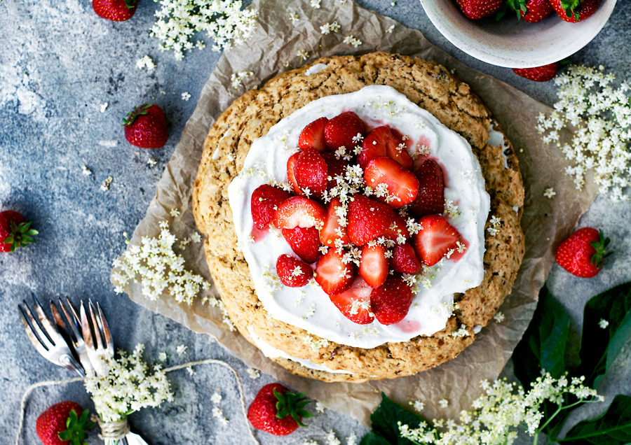 Giant scone cake layered with coconut whipped cream and strawberries, surrounded by elderflower, strawberries, and with a blue background