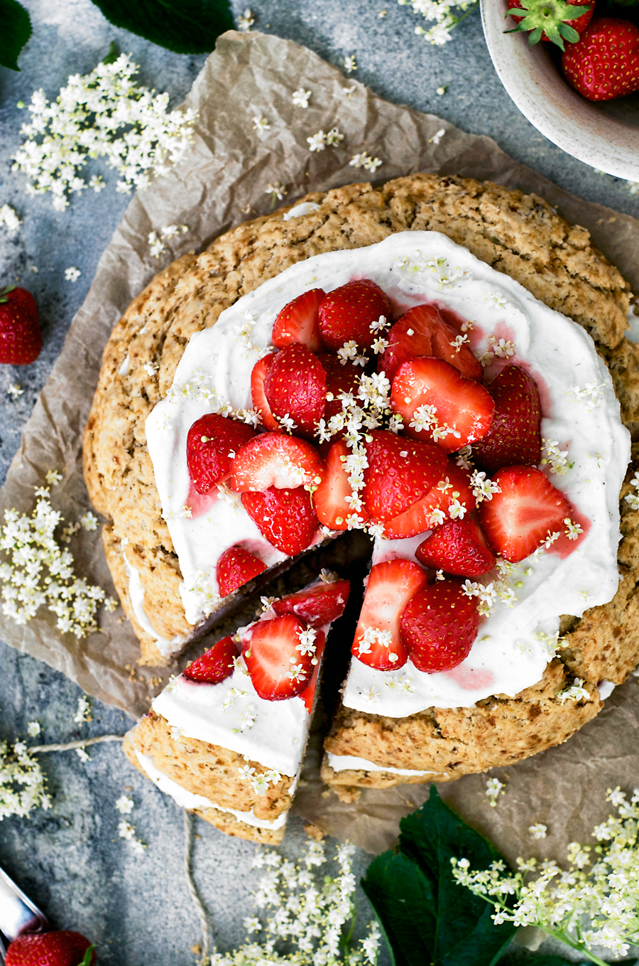Giant scone cake layered with coconut whipped cream and strawberries, surrounded by elderflower, strawberries, and with a blue background, one slice cut