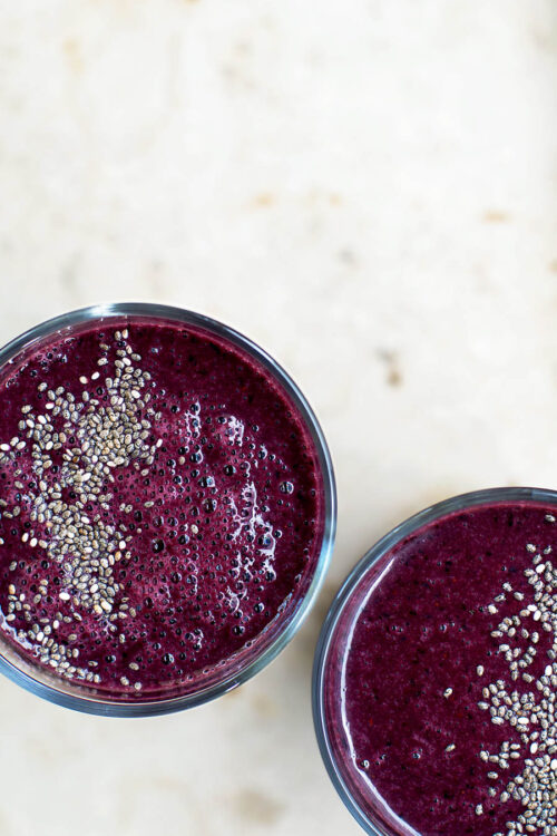 Two glasses filled with a dark purple blueberry smoothie, topped with chia seeds, top down view.