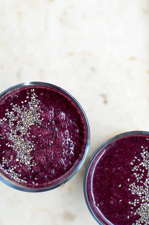 Two glasses filled with a dark purple blueberry smoothie, topped with chia seeds, top down view.
