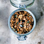 Olive oil granola with pumpkin seeds in a glass jar, top down view, on stone surface.