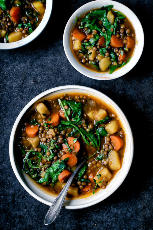 Three bowls of lentil potato stew with greens and carrots on a dark blue background.
