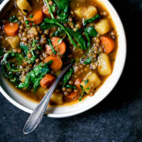 A bowl of lentil potato stew with greens and carrots on a dark blue background.