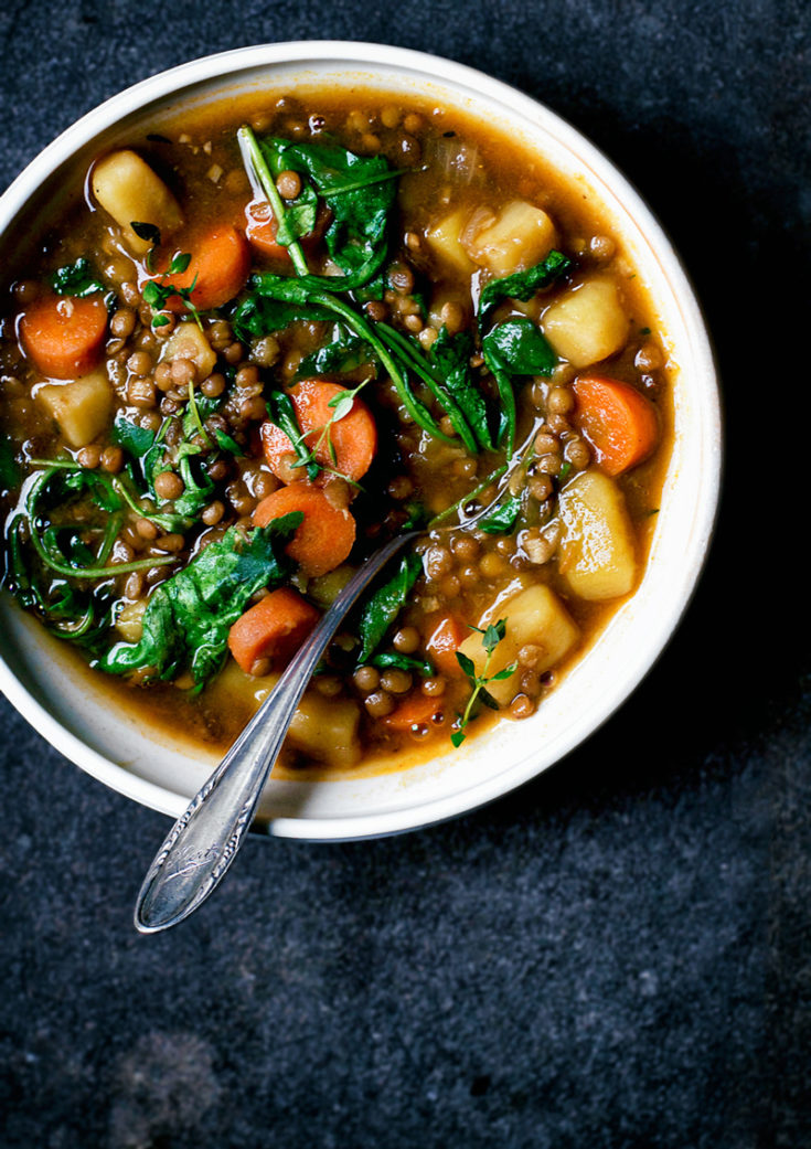 A bowl of lentil potato stew with greens and carrots on a dark blue background.