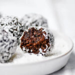 A chocolate date ball rolled in coconut on a small plate, with a bite taken out.