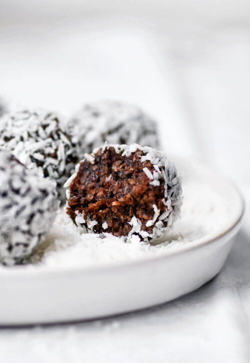 A chocolate date ball rolled in coconut on a small plate, with a bite taken out.