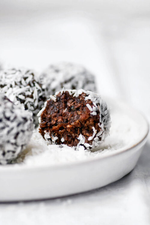 A chocolate date ball rolled in coconut on a small plate, with a bite taken out.