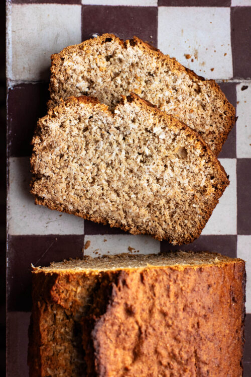 A loaf of coconut bread on a tile surface, top down view, with two slices cut.