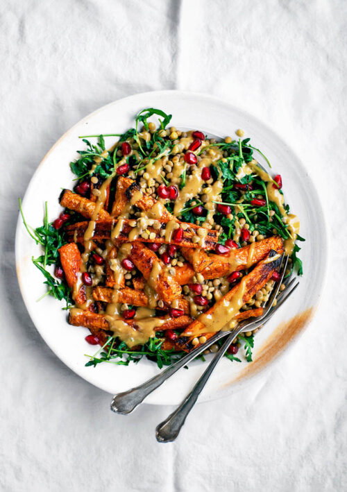 A plate full of roasted carrot salad with lentils, greens, and a tahini dressing on top.
