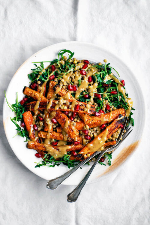 A plate full of roasted carrot salad with lentils, greens, and a tahini dressing on top.