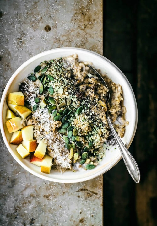 Apple porridge in a bowl on a ledge with fruit and seed toppings.