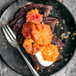 Three chocolate crepes on a plate with yogurt and blood orange slices.