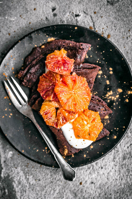 Three chocolate crepes on a plate with yogurt and blood orange slices.