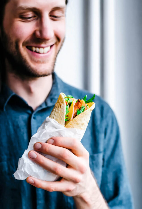 Man holding a wrap filled with vegetables and greens.