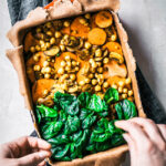 Hands adding spinach to cover sweet potato and chickpea filling in a pie.