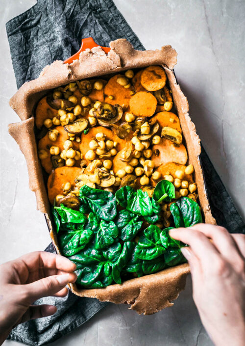 Hands adding spinach to cover sweet potato and chickpea filling in a pie.
