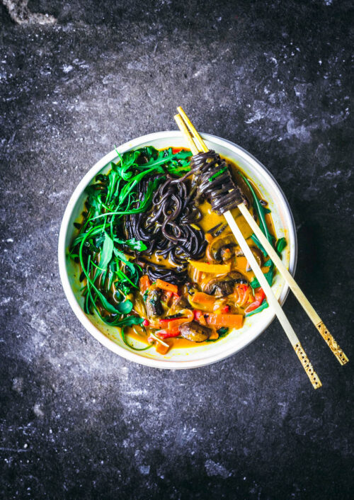 A vegetable noodle curry in a white bowl with chopsticks.