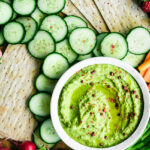 A small bowl of pea hummus with cucumber slices, radishes, crackers, and asparagus.