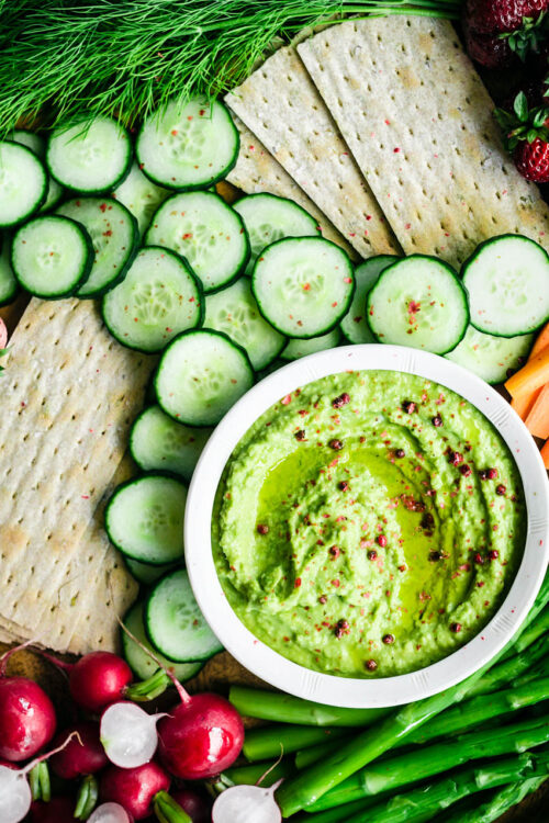 A small bowl of pea hummus with cucumber slices, radishes, crackers, and asparagus.