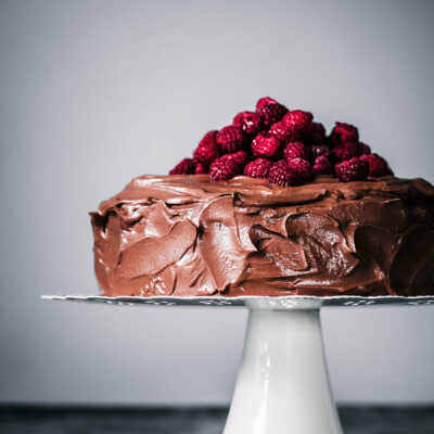 Chocolate ganache covered cake topped with fresh raspberries on a cake stand.