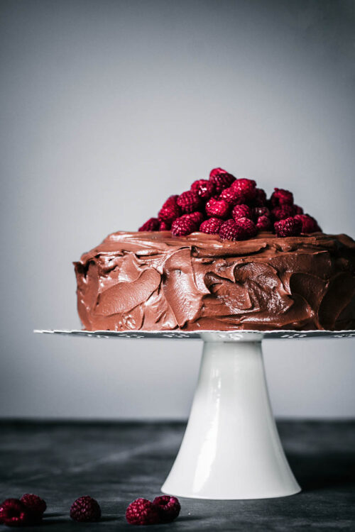 Chocolate ganache covered cake topped with fresh raspberries on a cake stand.