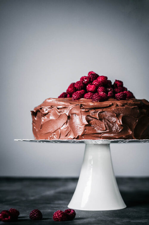 Chocolate ganache covered cake topped with fresh raspberries on a cake stand.