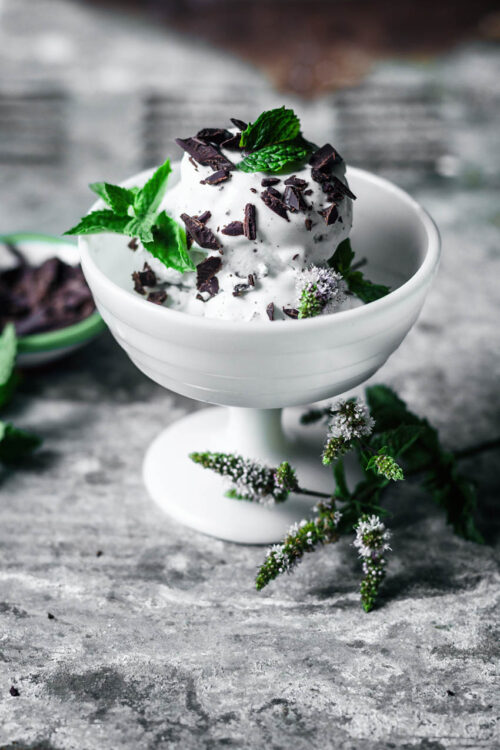 Vegan mint chocolate chip ice cream in a milk-glass coupe topped with more chocolate and mint leaves.