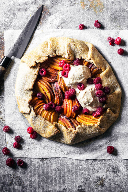 A peach and apricot galette with ice cream and raspberries on parchment paper.