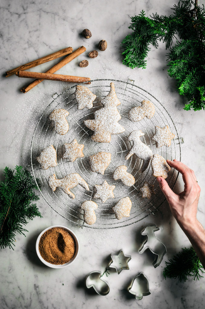 Spiced sugar cookie cut-outs shaped like trees, bells, mushrooms, and stars on a rack with spices and icing sugar.