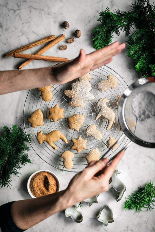 Spiced sugar cookie cut-outs shaped like trees, bells, mushrooms, and stars on a rack with spices and woman sprinkling icing sugar.
