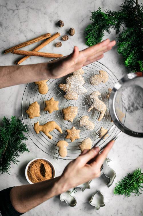 Spiced sugar cookie cut-outs shaped like trees, bells, mushrooms, and stars on a rack with spices and woman sprinkling icing sugar.