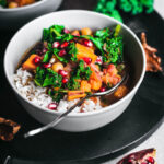 A bowl of chickpea, sweet potato, and kale soup with rice and pomegranate seeds.