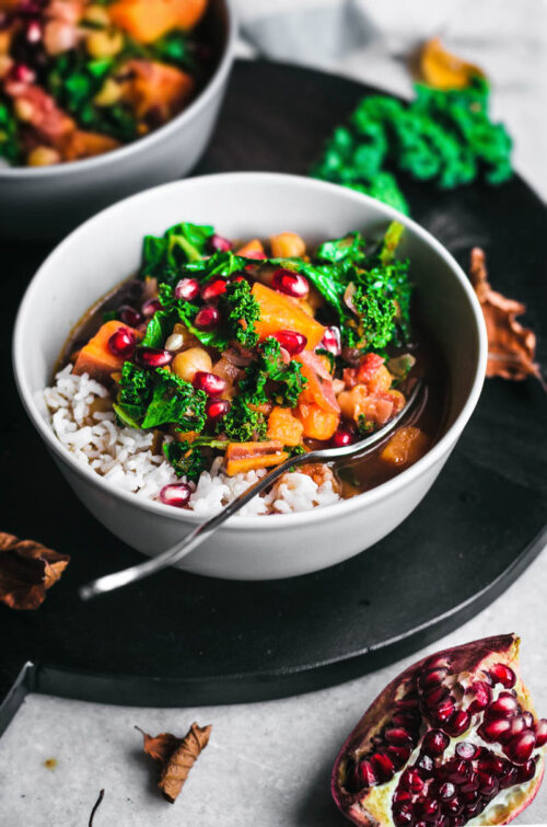 A bowl of chickpea, sweet potato, and kale soup with rice and pomegranate seeds.