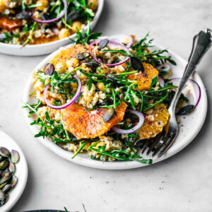 Two plates with grain and orange salad, with raw onion slices and greens.