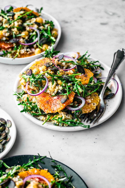 Two plates with grain and orange salad, with raw onion slices and greens.