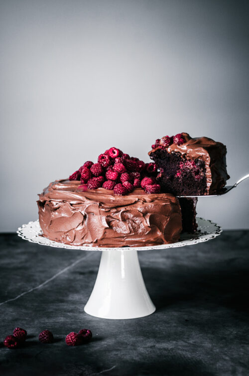 A large chocolate cake topped with raspberries, one slice being lifted out.