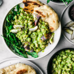 White bean salad with green sauce in bowl, top down view, surrounded by dishes of naan, more beans, shallots, and rucola