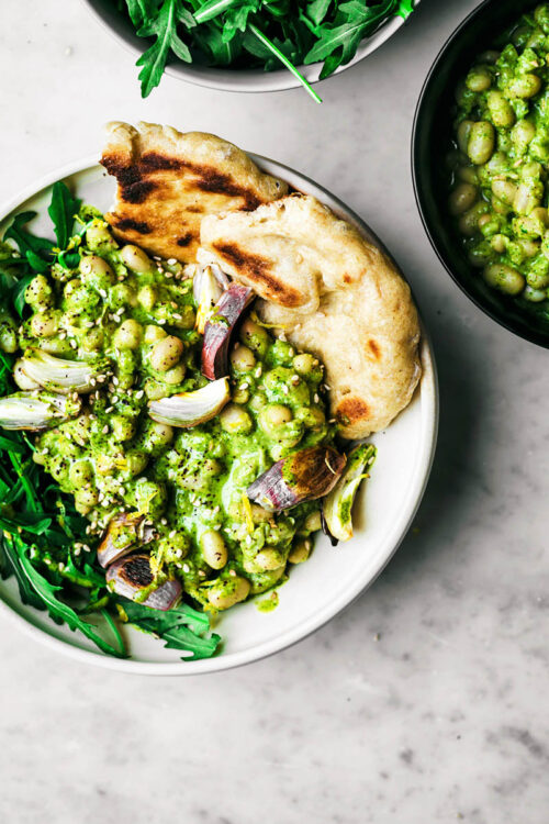 White bean salad with green sauce in a bowl with greens, naan, and cooked shallots.