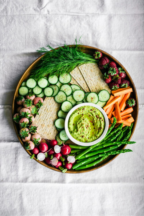 A large platter with green dip, vegetables, crackers, and berries.
