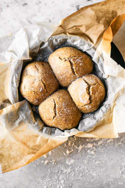 Four crusty buns with cracked tops in parchment lined cast iron pan, on marble surface