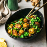 Golden-coloured potato salad with greens in black bowl, front view.