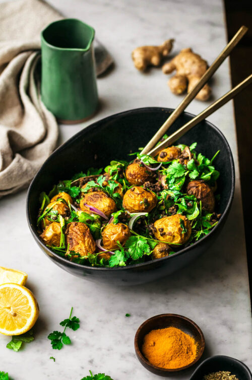 Golden-coloured potato salad with greens in black bowl, front view.