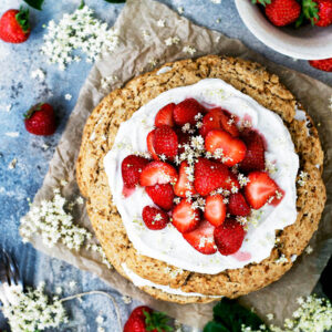 A large scone cake topped with whipped cream, strawberries, and elderflower blossoms.