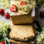 Top-front view of a loaf cake with two slices cut, topped with elderflowers and strawberries.