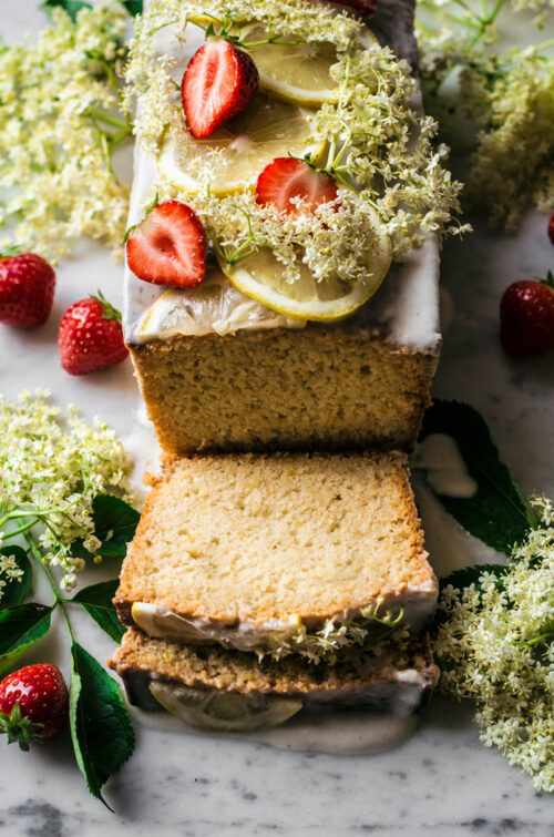 Top-front view of a loaf cake with two slices cut, topped with elderflowers and strawberries.