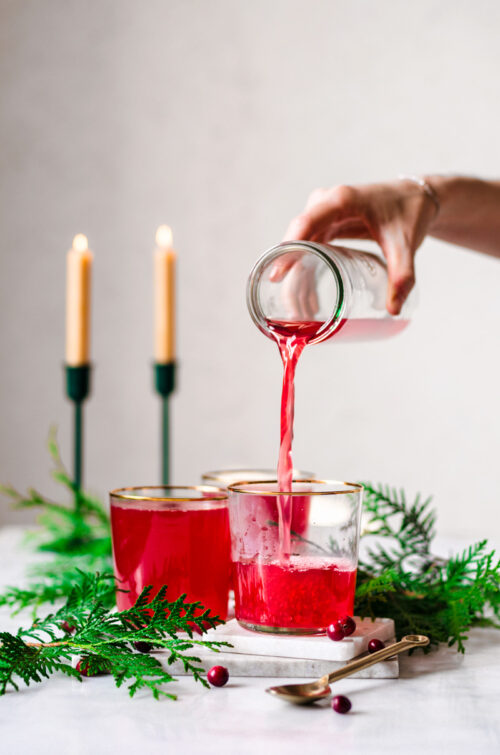 Hand pouring tall jar of cranberry tea into glass.