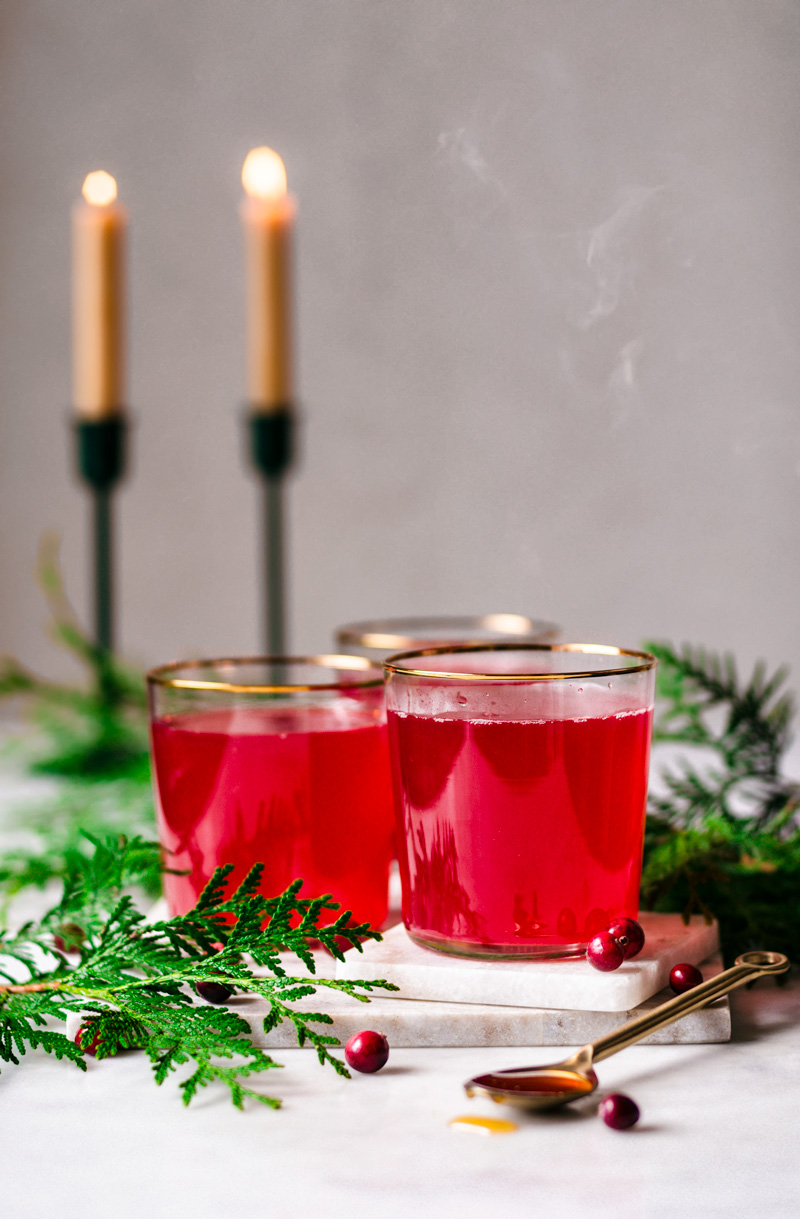 Three glasses of cranberry tea, steaming, with candles in the background.